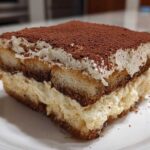 A close-up of a rich slice of tiramisu recipe, showing layers of coffee-soaked ladyfingers and creamy mascarpone, dusted with cocoa.