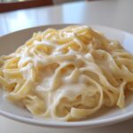 Close-up of fettuccine pasta generously coated in rich, creamy homemade alfredo sauce on a white plate.