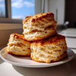 Three tall, flaky buttermilk biscuits stacked on a white plate, showing golden brown tops and layered interiors.
