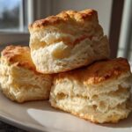 A close-up stack of three tall, flaky buttermilk biscuits with golden tops on a white plate.