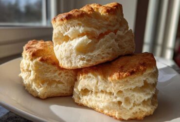 A close-up stack of three tall, flaky buttermilk biscuits with golden tops on a white plate.