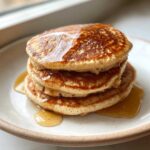 A close-up of three fluffy almond flour pancakes stacked high and drizzled generously with golden syrup.