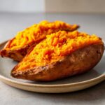 Two halves of a perfectly fluffy baked sweet potato, showing bright orange flesh, resting on a stoneware plate.