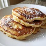 A close-up of a stack of fluffy banana pancakes dusted generously with powdered sugar on a white plate.