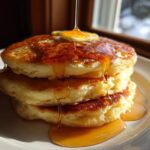 A tall stack of three fluffy buttermilk pancakes being drizzled with golden syrup and topped with melting butter.
