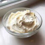 A clear glass bowl filled with freshly whipped, fluffy cream cheese frosting sitting on a white surface.