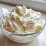 A clear glass bowl filled with freshly whipped, fluffy cream cheese frosting, showing soft peaks.