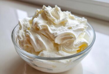 A clear glass bowl filled with freshly whipped, fluffy cream cheese frosting, showing soft peaks.