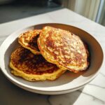Three golden brown greek yogurt pancakes stacked on a light ceramic plate, beautifully lit by sunlight.