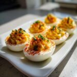 Close-up of six creamy deviled eggs garnished with paprika and fresh chives on a white platter.