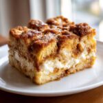 A close-up of a square slice of baked french toast casserole featuring a thick cream cheese layer.