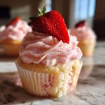 A close-up of a delicious strawberry cupcake topped with pink strawberry frosting and a fresh strawberry slice.
