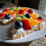 A close-up of a slice of fruit pizza sugar cookie topped with white frosting, blueberries, strawberries, kiwi, and orange segments.