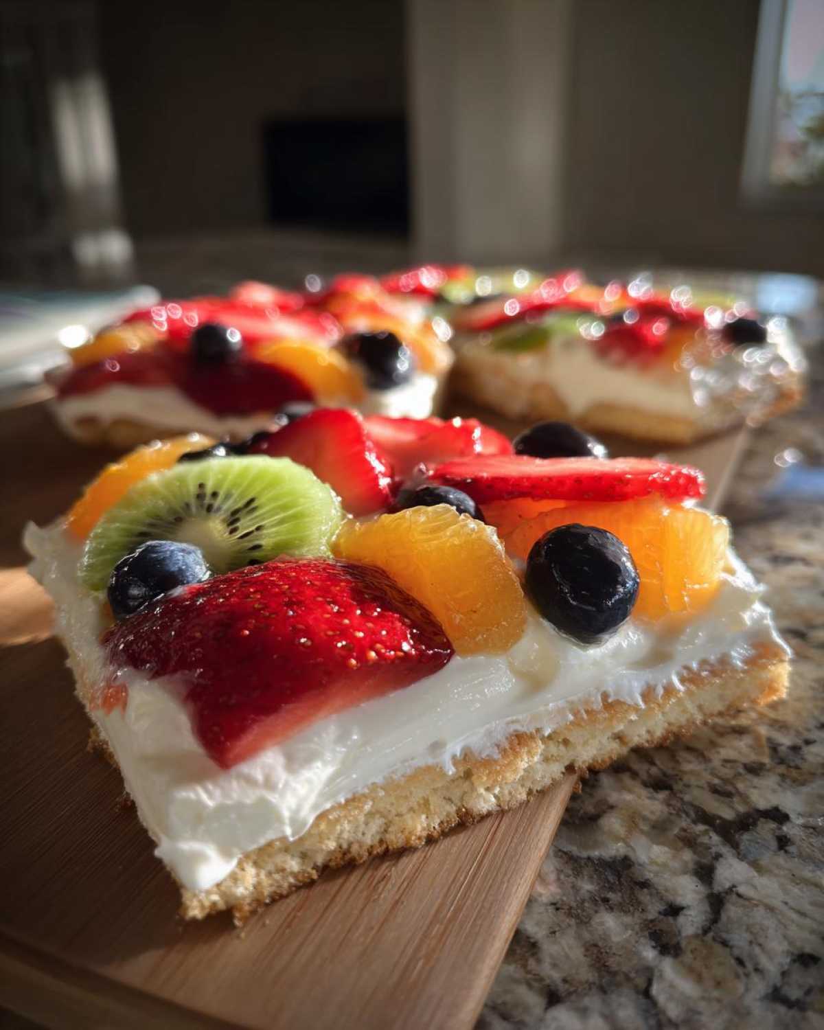 Close-up of a slice of fruit pizza sugar cookie topped with cream cheese frosting and fresh fruit like kiwi, strawberries, and blueberries.