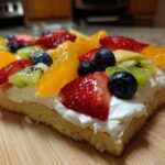 Close-up of a slice of fruit pizza sugar cookie topped with cream cheese frosting and fresh strawberries, blueberries, kiwi, and orange segments.