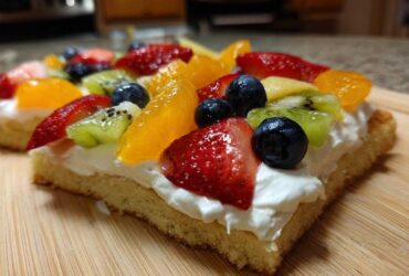 Close-up of a slice of fruit pizza sugar cookie topped with cream cheese frosting and fresh strawberries, blueberries, kiwi, and orange segments.