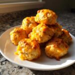 A stack of freshly baked, golden cream cheese sausage balls topped with melted cheddar cheese on a white plate.