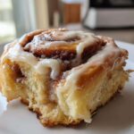 A single, soft, freshly baked cinnamon roll topped generously with white icing, resting on a white plate.