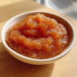 Close-up of chunky, caramelized homemade applesauce served in a small white bowl on a wooden surface.