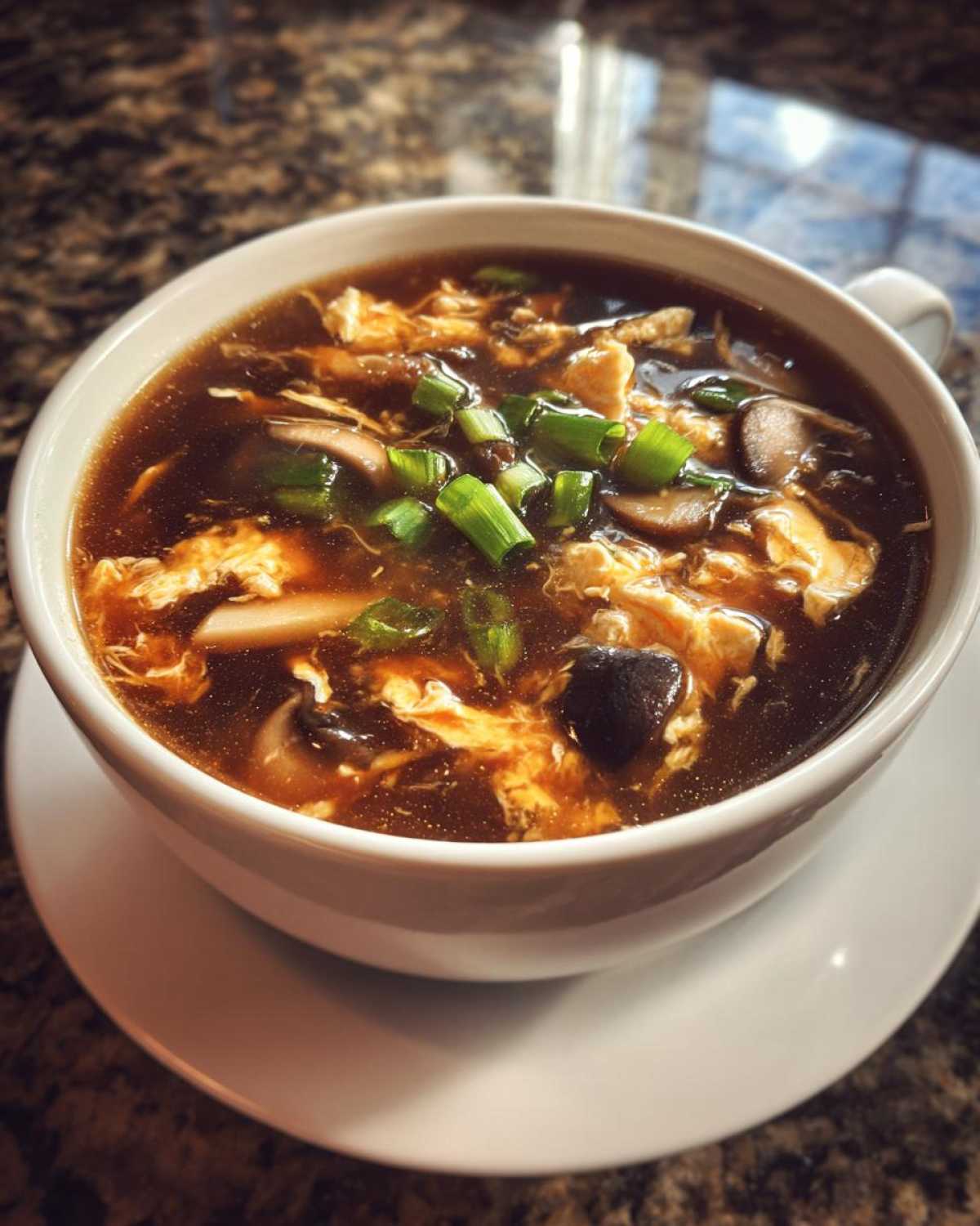 A close-up of a steaming bowl of hot and sour soup recipe, featuring dark broth, egg ribbons, mushrooms, and green onions.