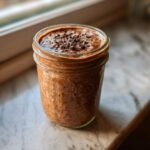 A close-up of chocolate overnight oats in a glass jar, topped with cacao nibs, sitting by a window.