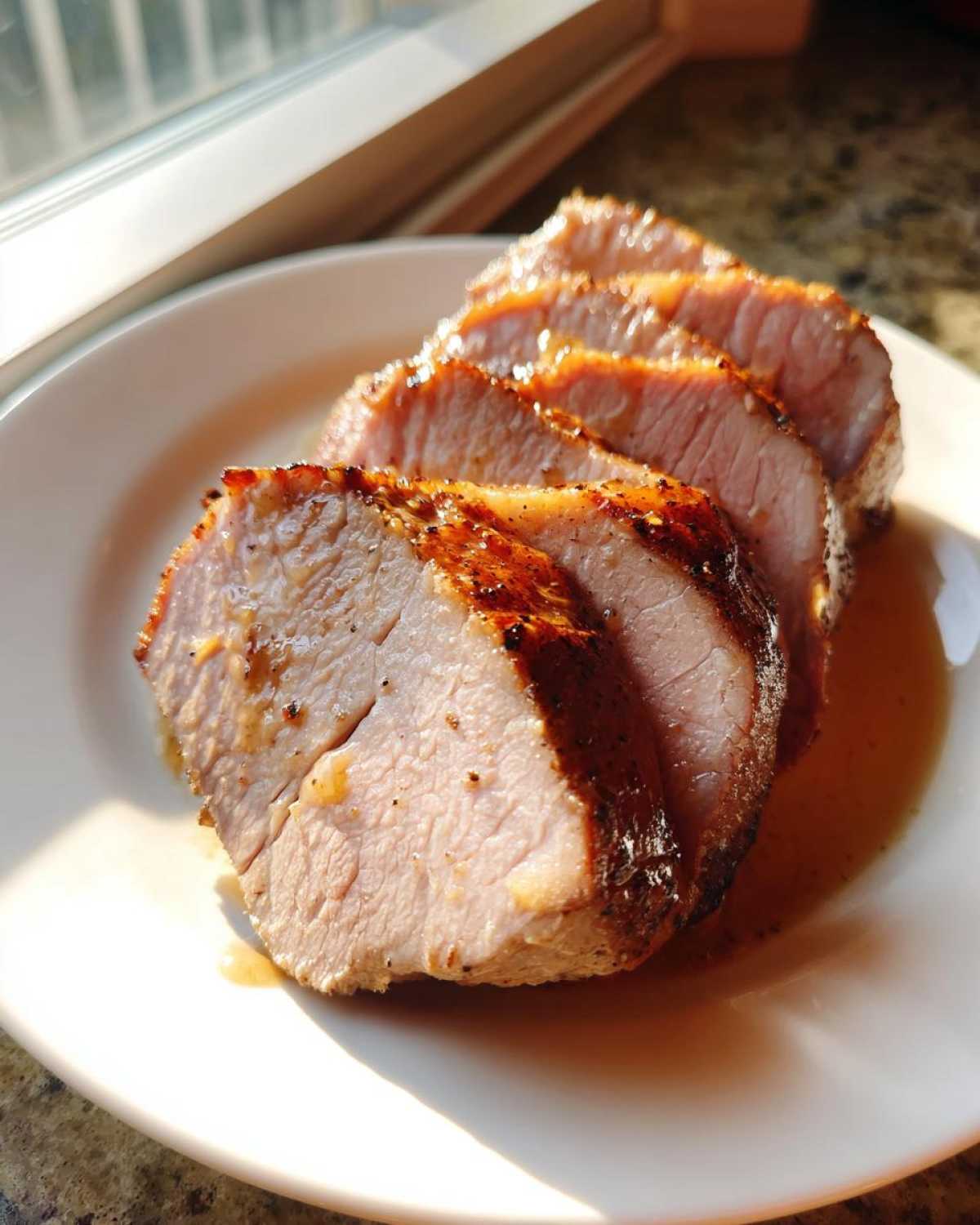 Close-up of four thick, juicy slices of pork loin roast resting in their pan juices on a white plate.