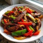 Close-up of juicy steak fajitas mixed with vibrant red and green bell peppers and onions on a white plate.