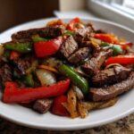Close-up of seasoned steak fajitas mixed with colorful sautéed red and green bell peppers and onions on a white plate.