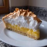 A close-up slice of lemon meringue pie showing a thick layer of bright yellow curd and tall, toasted meringue.