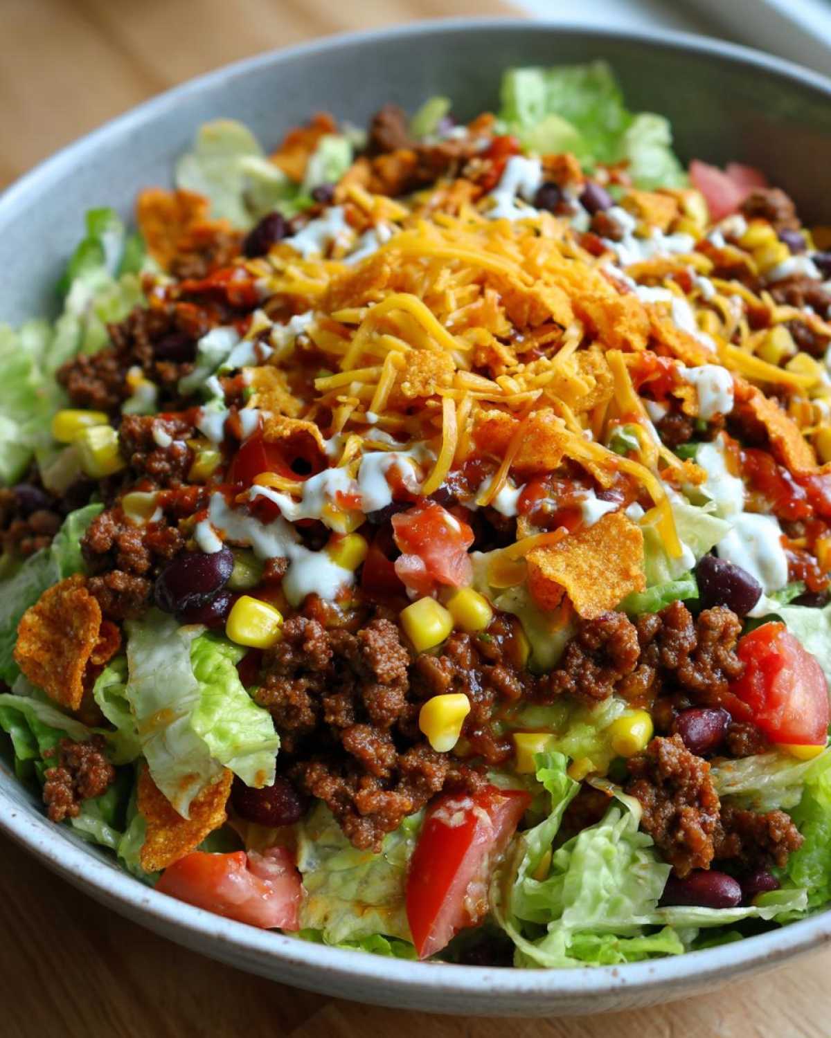 A close-up view of a hearty taco salad featuring seasoned ground beef, lettuce, corn, tomatoes, black beans, cheese, and sour cream drizzle.