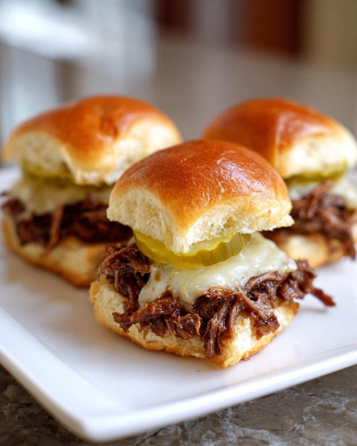 Three delicious Mississippi pot roast sliders featuring shredded beef, melted cheese, and a pickle slice on soft buns.