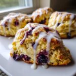 Close-up of a flaky blueberry scone topped with thick vanilla glaze dripping down the sides.