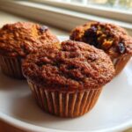 Close-up of three freshly baked, moist bran muffins with raisins, sitting on a white plate near a window.