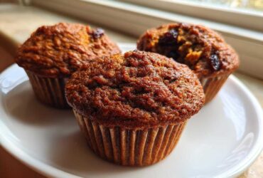 Close-up of three freshly baked, moist bran muffins with raisins, sitting on a white plate near a window.