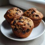 Three freshly baked, moist bran muffins studded with raisins sitting on a small white plate.