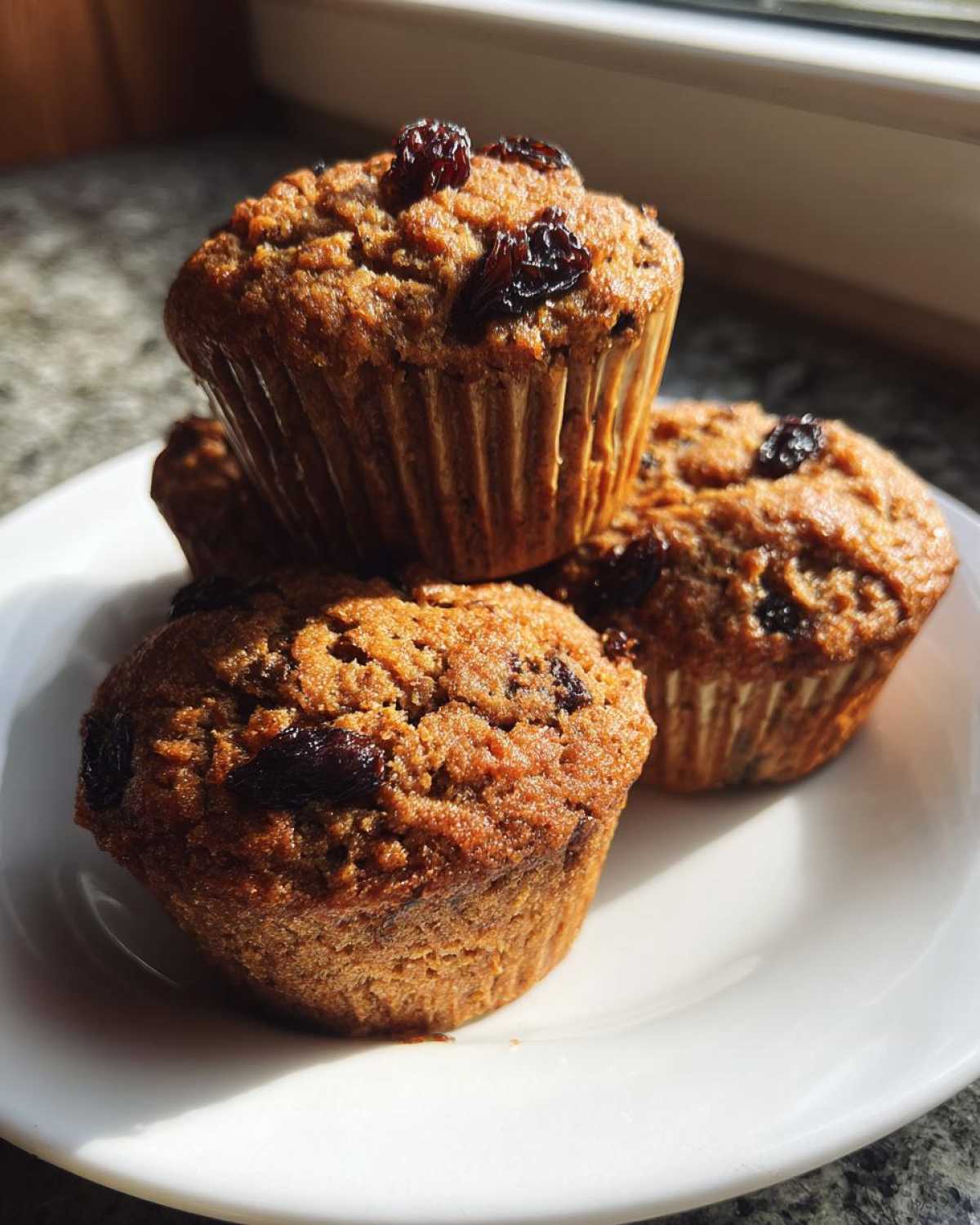 A stack of four moist bran muffins topped with dark raisins, sitting on a white plate in bright sunlight.