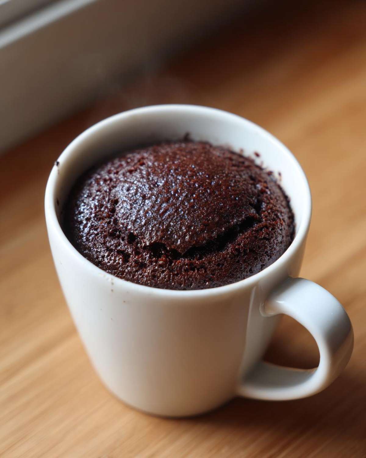Close-up of a moist, dark chocolate mug cake rising over the rim of a white ceramic mug.