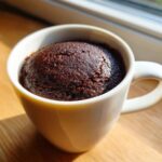 A moist, dark chocolate mug cake risen above the rim of a white ceramic mug, sitting on a wooden surface.