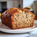 A golden-brown loaf of moist sugar free banana bread resting on a white plate.