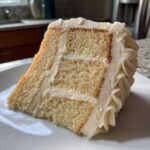 A close-up of a perfectly layered slice of moist vanilla cake recipe with thick white vanilla buttercream frosting.