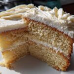 Close-up of a moist slice of vanilla cake recipe with thick white frosting layers.