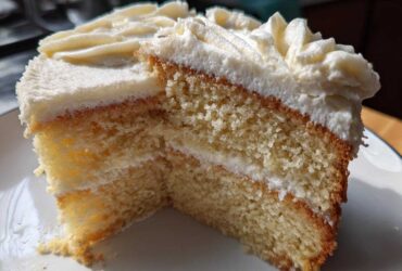 Close-up of a moist slice of vanilla cake recipe with thick white frosting layers.