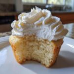 Close-up of a moist vanilla cupcakes with fluffy white frosting, showing the soft crumb after a bite was taken.