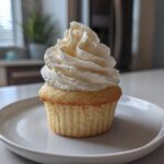 A single, moist vanilla cupcake topped with a tall swirl of white vanilla frosting on a white plate.