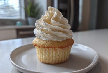 A single, moist vanilla cupcake topped with a tall swirl of white vanilla frosting on a white plate.
