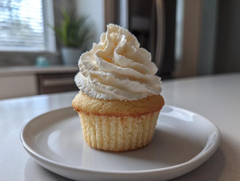 A single, moist vanilla cupcake topped with a tall swirl of white vanilla frosting on a white plate.