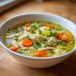 Close-up of a white bowl filled with steaming penicillin soup, featuring shredded chicken, sliced carrots, and fresh parsley garnish.