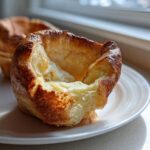 Close-up of a golden brown, perfectly risen Yorkshire pudding with a hollow center, sitting on a white plate.