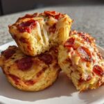Three freshly baked pizza muffins stacked on a white plate, one broken open showing a dramatic cheese pull.