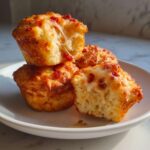 Four golden brown pizza muffins stacked on a white plate, showing a dramatic cheese pull from the broken muffin.